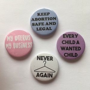 May include: Four round buttons with different messages printed on them. The buttons are pink, blue, purple, and white. The messages are "Keep Abortion Safe and Legal", "My Uterus My Business", "Every Child a Wanted Child", and "Never Again" with a hanger graphic.