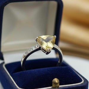 Könnte beinhalten: Ein silberner Ring mit einem großen, dreieckigen gelben Edelstein, präsentiert in einer blauen Samt-Ringbox. Der Ring ist mit kleinen, klaren Akzentsteinen besetzt. Die Box ist geöffnet und zeigt den Ring vor einem weichen, goldenen Hintergrund.