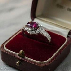 Op de afbeelding: Een zilveren ring met een grote ovale robijn edelsteen, omringd door kleinere diamanten, ligt in een vintage bordeauxrode sieradendoos met een rode fluwelen binnenkant. De ring heeft kleine, rechthoekige diamanten.