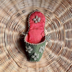 May include: A small, green fabric pouch with a red lining and silver bead embellishments. The pouch is decorated with floral patterns and is sitting on a woven surface.