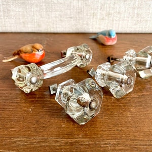 May include: Four clear glass cabinet knobs with a faceted design. The knobs have a silver metal base and are arranged on a wooden surface.