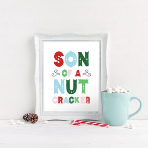 May include: A white framed print with the text "SON OF A NUT CRACKER" in colorful letters. The letters are in red, green, blue, and pink. The print is sitting on a white surface with a mug of hot chocolate and a candy cane.