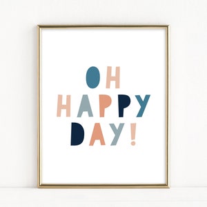 May include: A framed art print with the words "OH HAPPY DAY!" in a playful, colorful font. The text is in shades of blue, peach, and gray against a white background, framed in gold.