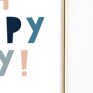 May include: A framed print with the words "HAPPY DAY!" in a playful, colorful font. The letters are in shades of peach, navy blue, and teal against a white background. The frame is gold.