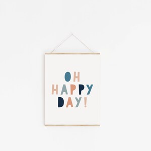 May include: A framed print with the words "OH HAPPY DAY!" in a playful, colorful font. The text is in shades of blue, coral, and peach against a white background. The frame is a light wood color, and the print is hanging on a white wall.