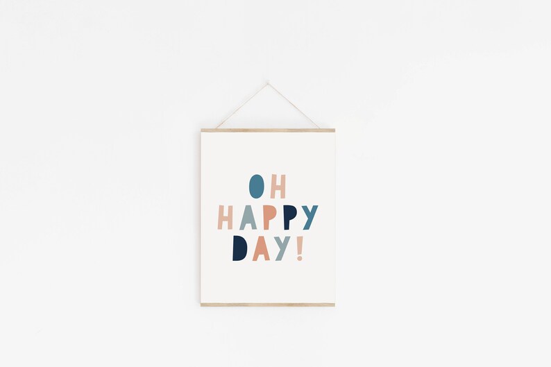 May include: A framed print with the words "OH HAPPY DAY!" in a playful, colorful font. The text is in shades of blue, coral, and peach against a white background. The frame is a light wood color, and the print is hanging on a white wall.