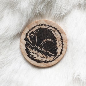 May include: A wooden circle with a black and white illustration of a curled up animal with a long tail. The animal is surrounded by a ring of small, black lines.