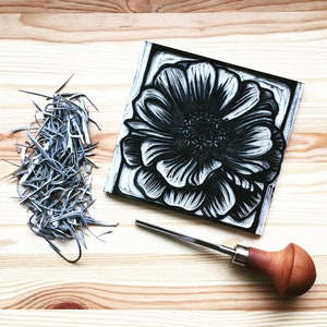 May include: A black and white woodblock print of a large flower with intricate details. A wood carving tool with a wooden handle is also pictured.