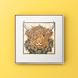 Puede incluir: Una impresi&oacute;n en madera de una vaca de las Tierras Altas con una corona de flores alrededor del cuello. La impresi&oacute;n es en tonos de marr&oacute;n, amarillo y verde, y est&aacute; enmarcada en un marco de madera claro.