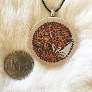 May include: A wooden pendant necklace with a hand-painted image of a curled up fox with orange and brown fur. The pendant is round and hangs from a black cord.