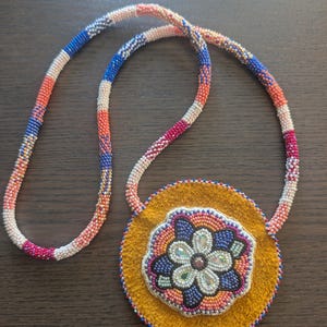 May include: A handmade necklace with a circular pendant displaying a beaded floral pattern on a yellow suede base. The necklace cord is made of colourful beads in red, white, blue, and orange. The pendant is approximately 10 cm in diameter.