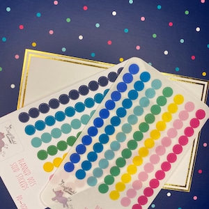 May include: Two sheets of planner dot stickers. One sheet features solid dots in shades of blue, green, and yellow. The other sheet has transparent dots in a rainbow of colors. The stickers are on a navy blue background with white and gold accents.