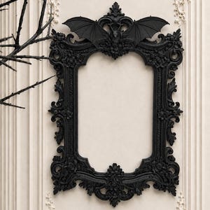 May include: A rectangular black picture frame featuring ornate, intricate scrollwork and floral carvings. The top centre of the frame is adorned with a detailed sculpted bat figure with its wings fully extended across the upper border.