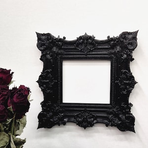 Puede incluir: Un marco de fotos negro ornamentado con un centro blanco vacío cuelga de una pared blanca. El marco presenta motivos florales y de volutas. Un ramo de rosas rojas secas está en un jarrón negro a la izquierda del marco.