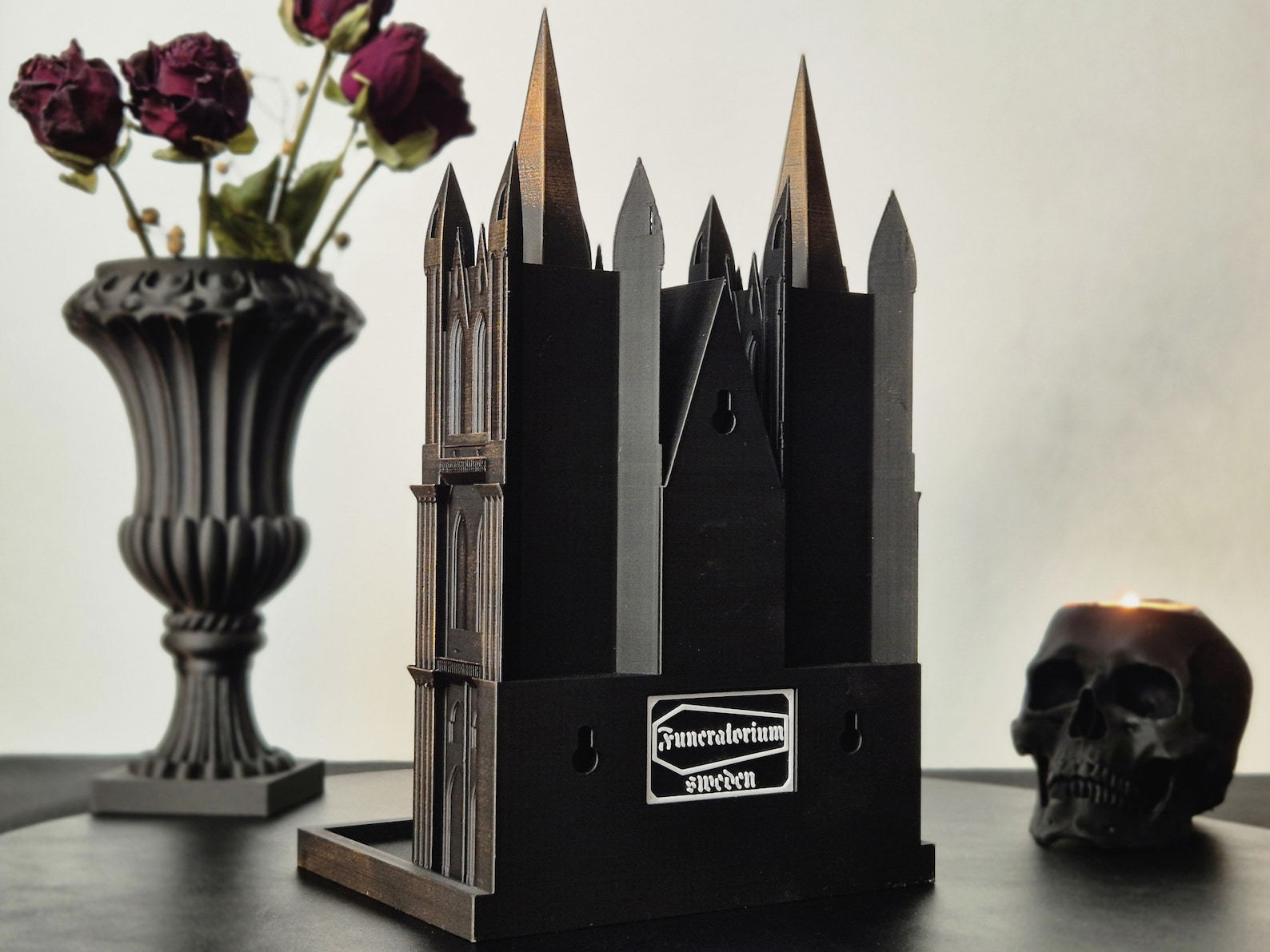 Back view of a black and gold Gothic cathedral shelf with mounting holes, displayed beside a black urn with dried roses and a black skull candle