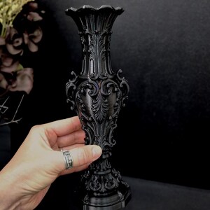 May include: A matte black vase featuring an intricate, raised relief pattern with ornate scrollwork and acanthus leaf motifs. The vase has a flared, scalloped rim, a slender neck, a bulbous body, and a wide, decorative pedestal base.