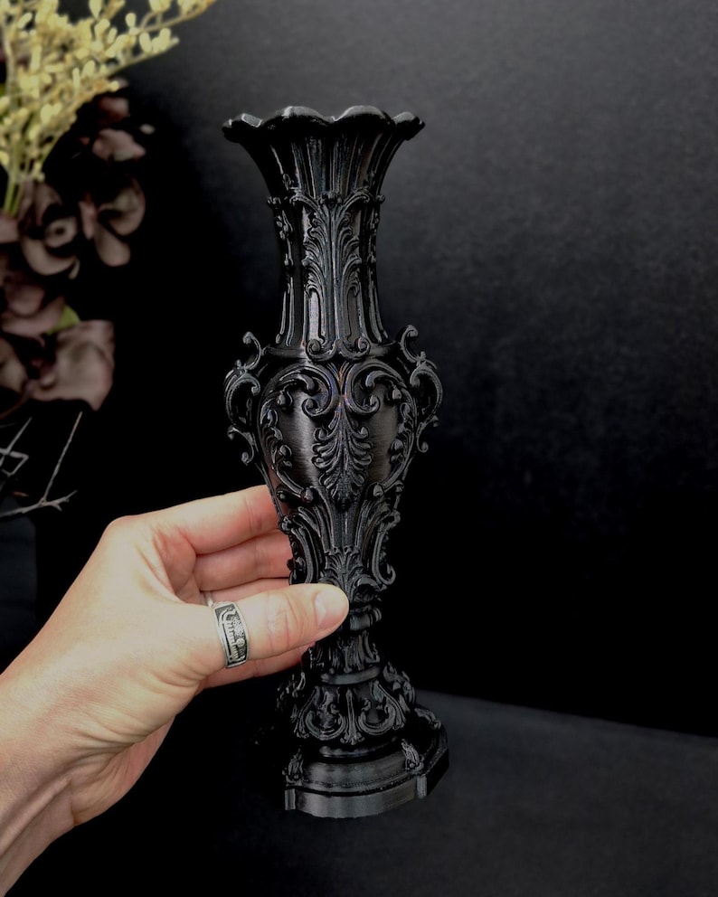 May include: A matte black vase featuring an intricate, raised relief pattern with ornate scrollwork and acanthus leaf motifs. The vase has a flared, scalloped rim, a slender neck, a bulbous body, and a wide, decorative pedestal base.