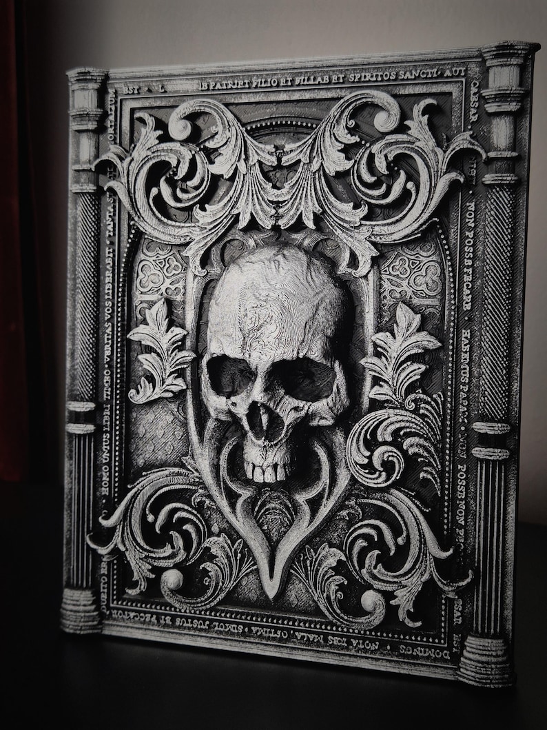 May include: A black and white 3D printed book cover with an ornate design featuring a skull in the center. The cover has a gothic style with intricate details and a textured surface. The text on the cover reads "DOMINUS NOTA TOBS MALA, OFTIMA SUMOL JUSTUS BT PECCATOR", "UBITO BRA", "HOMO UNTUS LIBIU TIMRO VERITAS VOS LIBERABIT", "TANTAST", "CORTALIUM", "CABSAR", "NTT NON POSSE FECARE", "HABEMUS PAPARC NON POSSE", "NON PH", "SAR BST", "STAL", "FIS PATRIET PILIO ET VILLAB ET SPIRITOS SANCTI AUT".