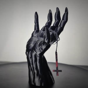 Może przedstawiać: Czarna, drukowana w 3D rzeźba dłoni z długimi, spiczastymi paznokciami. Czarny sznurek z czerwono-czarnym wisiorkiem z odwróconym krzyżem wisi z dłoni. Rzeźba jest wystawiona na ciemnej powierzchni.