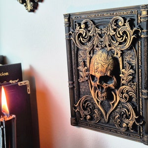 May include: A black and gold decorative wall plaque with a skull in the center. The skull is surrounded by ornate, gold-painted scrollwork. The plaque is framed with a black border and has a raised, textured surface.
