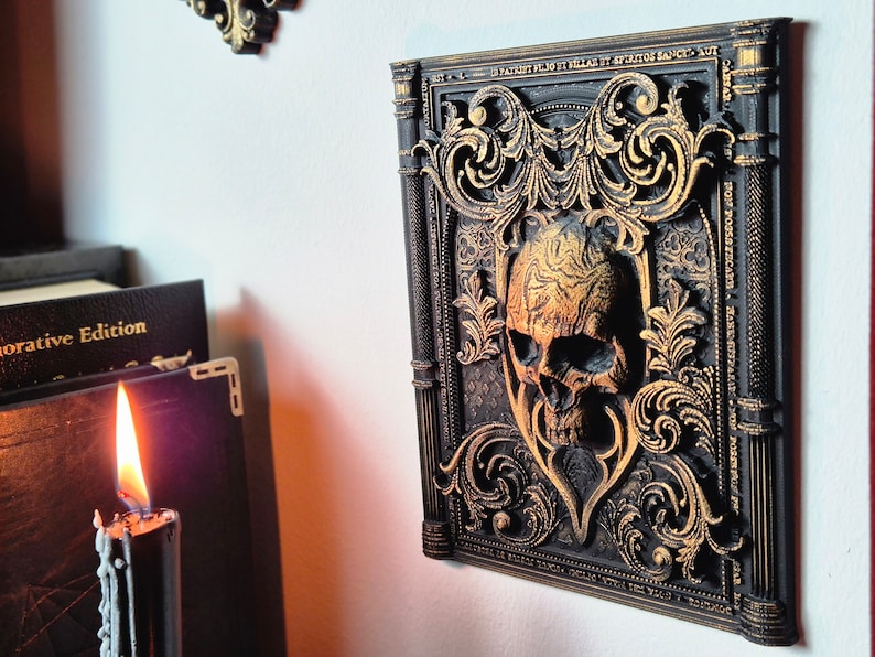 May include: A black and gold decorative wall plaque with a skull in the center. The skull is surrounded by ornate, gold-painted scrollwork. The plaque is framed with a black border and has a raised, textured surface.