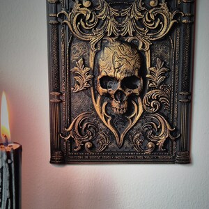 May include: A black and gold framed wall decor with a 3D skull in the center. The frame is ornate with gold accents and the skull is textured with gold paint.