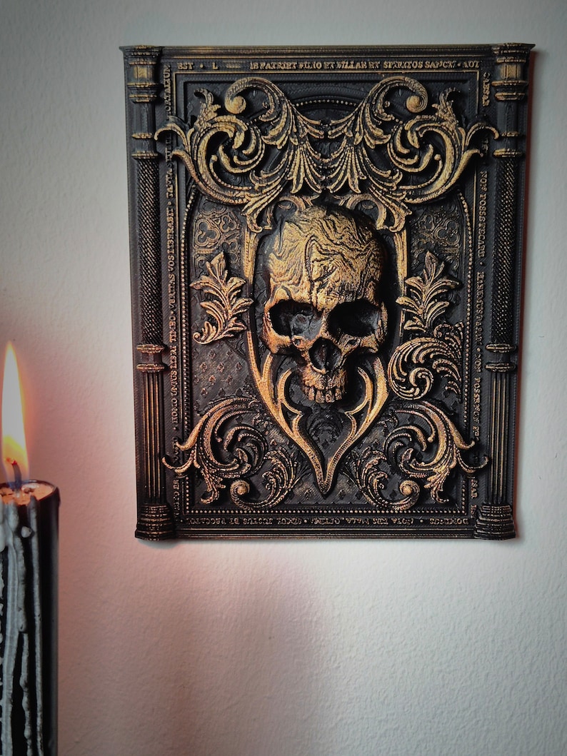 May include: A black and gold framed wall decor with a 3D skull in the center. The frame is ornate with gold accents and the skull is textured with gold paint.