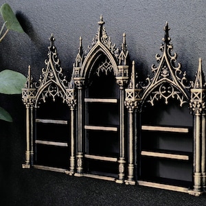 Könnte beinhalten: Schwarzes und goldenes Wandregal im gotischen Stil mit drei Abschnitten. Das Regal weist aufwendige Details auf, darunter Spitzbögen und dekorative Türme. Die Regale sind aus Holz und eignen sich ideal zum Ausstellen kleiner Gegenstände.