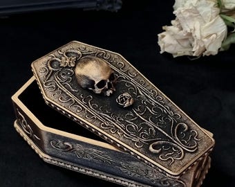 Può includere: Una scatola a forma di bara con finitura bronzo metallizzato e dettagli neri. Il coperchio presenta un teschio in rilievo e una piccola rosa, circondati da intricati motivi a volute. I lati mostrano disegni decorativi in rilievo.