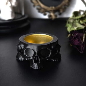 Pode incluir: Um castiçal preto em forma de caveira com interior dourado. O castiçal é feito de um material escuro e tem um design detalhado de caveira. O produto é um item decorativo.
