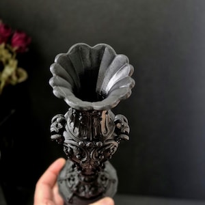 May include: A matte black vase featuring an ornate, relief-carved surface with intricate scrollwork and floral motifs. The vessel has a wide, scalloped rim and a tapered neck that expands into a detailed, textured body with a circular base.