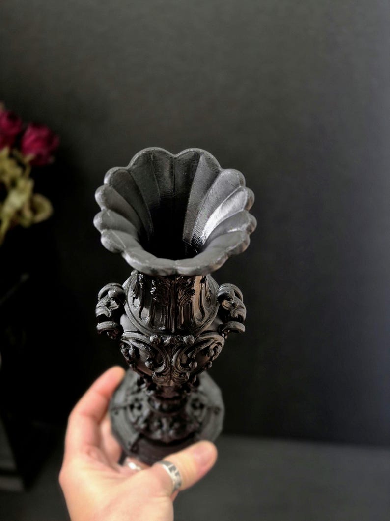 May include: A matte black vase featuring an ornate, relief-carved surface with intricate scrollwork and floral motifs. The vessel has a wide, scalloped rim and a tapered neck that expands into a detailed, textured body with a circular base.