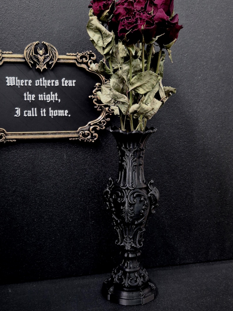 May include: A black ornate vase with intricate relief patterns holds a bouquet of dried dark red roses. Beside it, a black rectangular plaque with gold trim and a bat emblem displays the white text: Where others fear the night, I call it home.