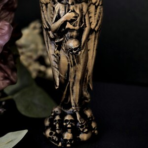 May include: A bronze-colored angel statue with large, detailed wings. The angel holds a skull in one hand and rests the other on its chest. The base of the statue is adorned with multiple skulls. The statue is set against a dark background.