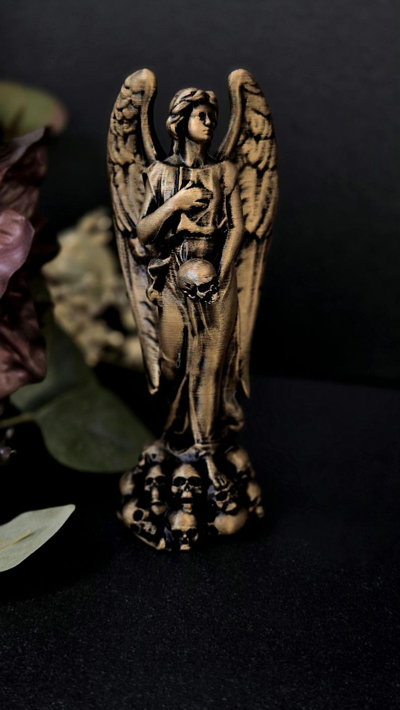 May include: A bronze-colored angel statue with large, detailed wings. The angel holds a skull in one hand and rests the other on its chest. The base of the statue is adorned with multiple skulls. The statue is set against a dark background.