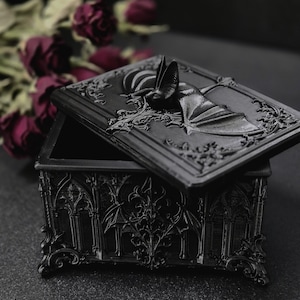 Könnte beinhalten: Eine mattschwarze rechteckige Schatulle mit einem Deckel, der in der Mitte eine skulptierte Fledermaus zeigt. Die Schatulle ist an allen Seiten mit detaillierten Reliefs aus gotischen Bögen und kunstvollen Ornamenten verziert.