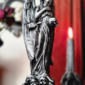 May include: A silver-colored angel statue holding a skull, standing atop a base of skulls. The angel has detailed wings and flowing robes. A candle and a framed picture are in the blurred background.