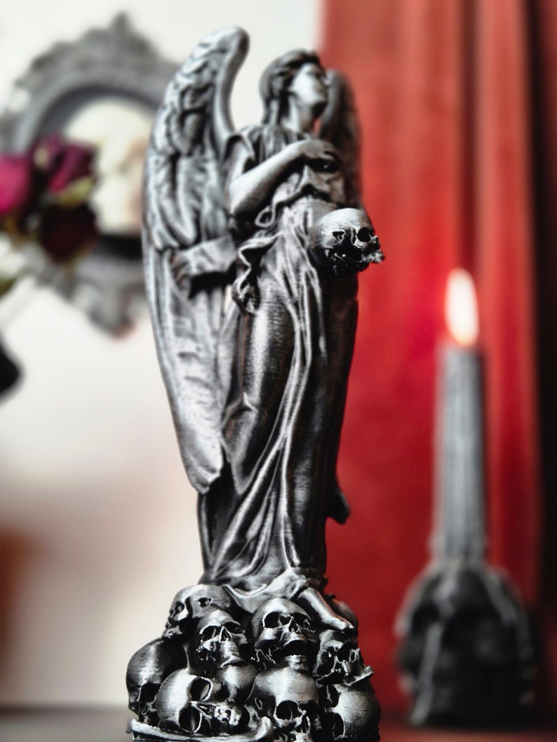 May include: A silver-colored angel statue holding a skull, standing atop a base of skulls. The angel has detailed wings and flowing robes. A candle and a framed picture are in the blurred background.
