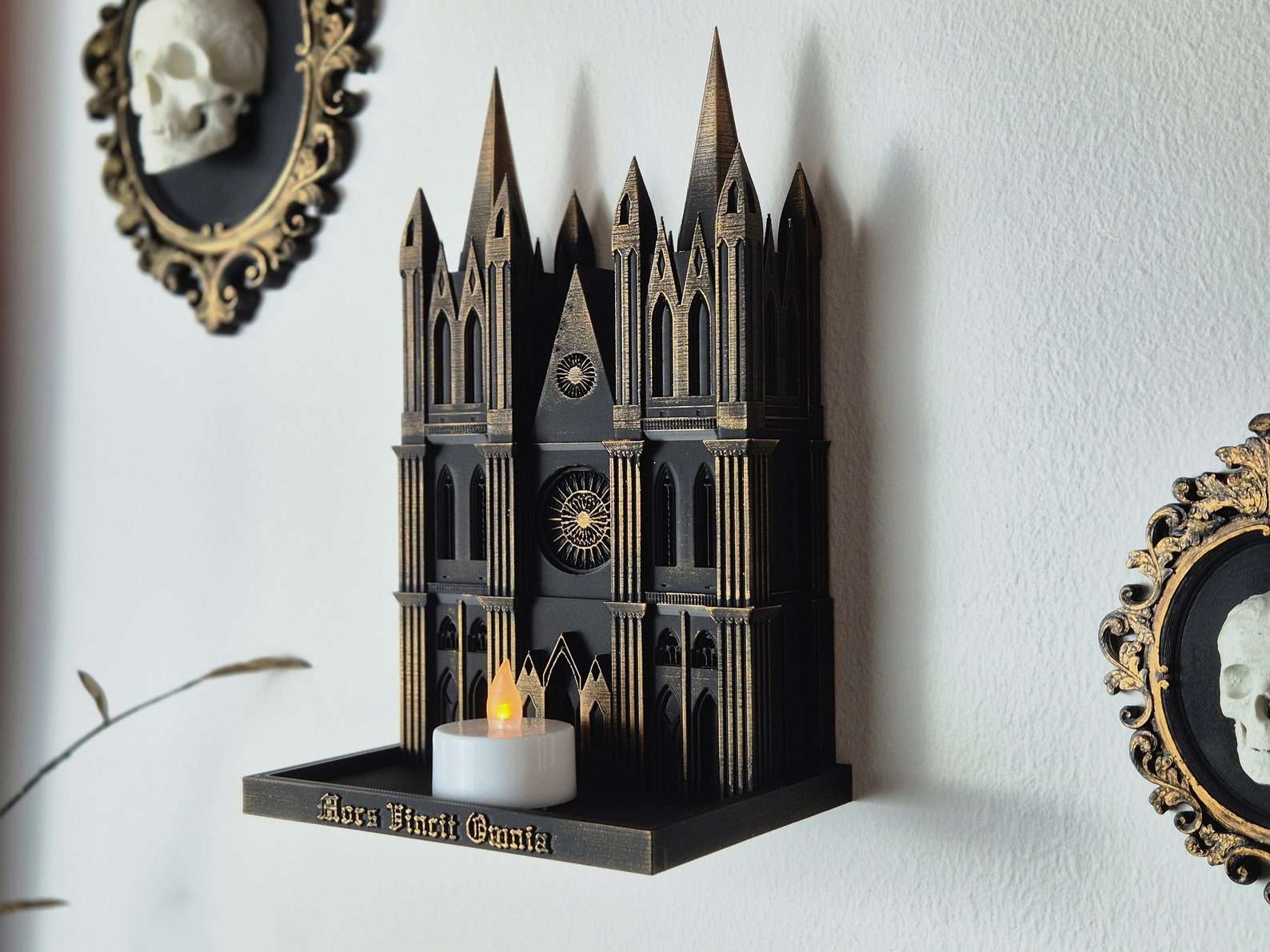 Black and gold Gothic cathedral wall shelf with LED tealight candle, mounted on a white wall between two framed skull cameos