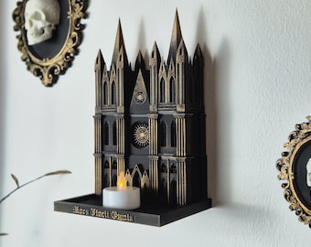 May include: A black and gold 3D printed gothic cathedral wall decor with a small candle on a shelf. The shelf has the inscription "Mora Vincit Omnia".