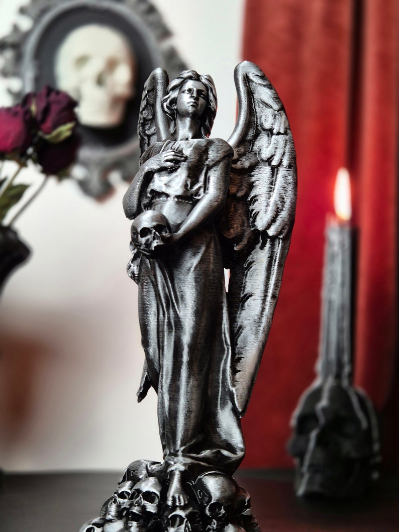 May include: A silver-colored angel statue holding a skull, with detailed wings and a flowing gown. The statue is standing on a base of skulls. A lit candle and a framed skull are in the background, creating a gothic theme.