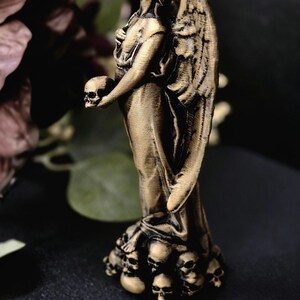 May include: A bronze-colored angel statue holding a skull. The angel has large detailed wings and stands atop a base of small skulls. The statue is set against a dark background with hints of foliage.