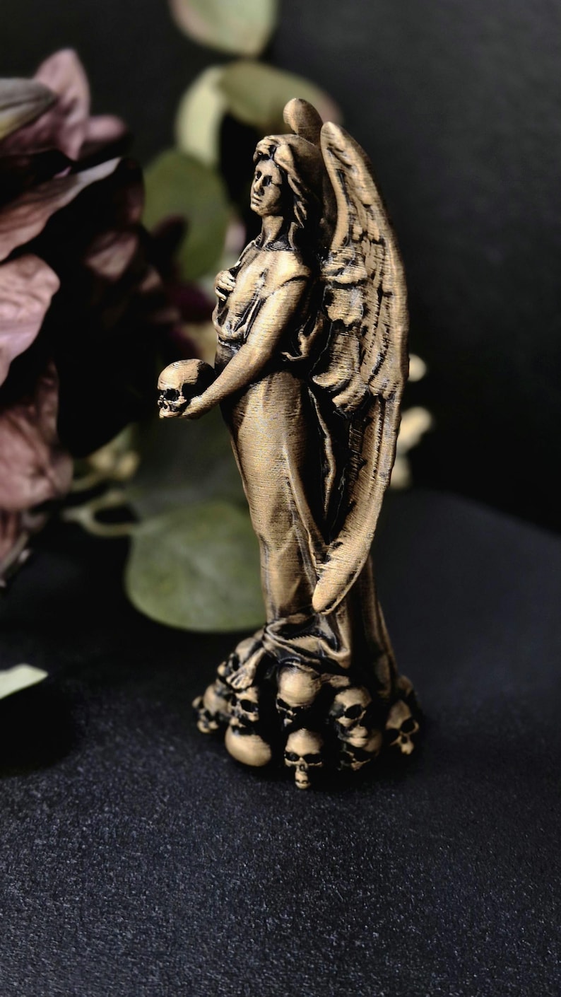 May include: A bronze-colored angel statue holding a skull. The angel has large detailed wings and stands atop a base of small skulls. The statue is set against a dark background with hints of foliage.