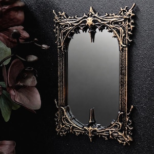 May include: Ornate black and gold mirror with an arched top and intricate, gothic-inspired detailing. The mirror has a rectangular shape and is mounted on a dark textured wall. The frame features a spiky, decorative design.