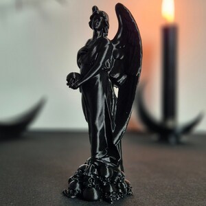May include: A black statue of an angel holding a skull, with large wings and a flowing gown. The statue is perched atop a base of skulls. A lit candle and a crescent moon shape are in the background.