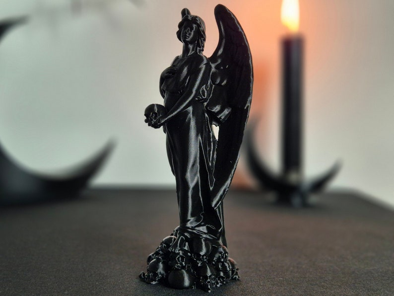 May include: A black statue of an angel holding a skull, with large wings and a flowing gown. The statue is perched atop a base of skulls. A lit candle and a crescent moon shape are in the background.