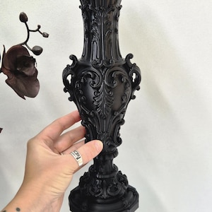 May include: A matte black vase featuring an ornate, raised relief design with intricate scrollwork and leaf motifs. The vase has a flared, scalloped rim, a slender neck, a bulbous body, and a wide, decorative pedestal base with detailed carvings.