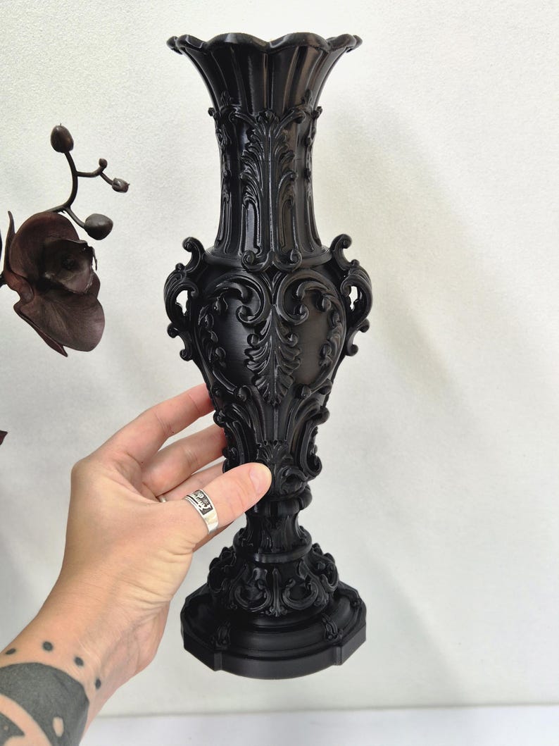 May include: A matte black vase featuring an ornate, raised relief design with intricate scrollwork and leaf motifs. The vase has a flared, scalloped rim, a slender neck, a bulbous body, and a wide, decorative pedestal base with detailed carvings.