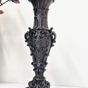 May include: A tall, matte black vase featuring an intricate, raised relief design with ornate scrollwork, acanthus leaf motifs, and a scalloped rim. The vessel has a slender neck, a bulbous body, and a wide, decorative pedestal base.