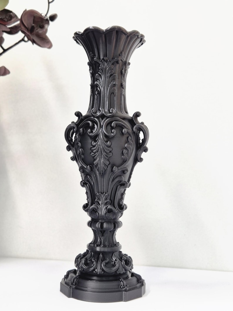 May include: A tall, matte black vase featuring an intricate, raised relief design with ornate scrollwork, acanthus leaf motifs, and a scalloped rim. The vessel has a slender neck, a bulbous body, and a wide, decorative pedestal base.